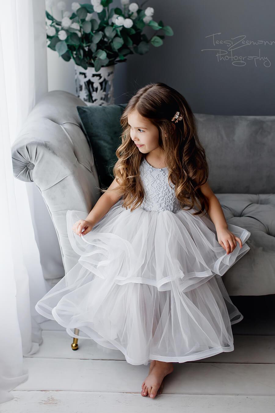 A little girl is sitting on the couch wearing a grey tulle dress. She has brown hair