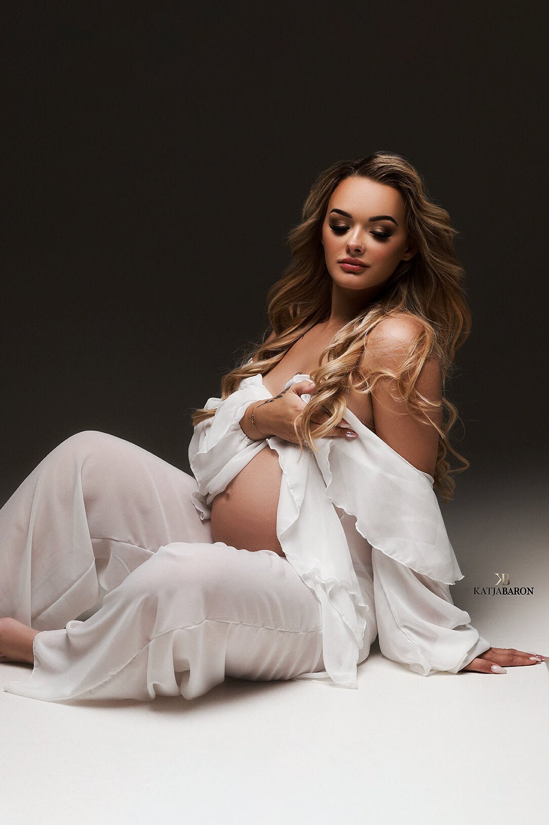 Blond pregnant model with long hair has her eyes closed while sitting on thew studio floor during her maternity photoshoot. She wears a maternity chiffon set of a short robe and pants. She holds her breast covering the body with the robe.