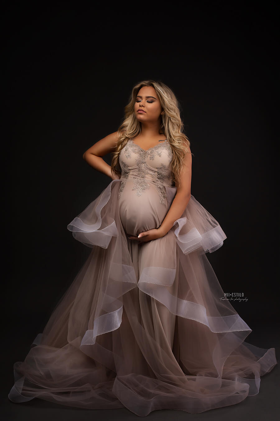Pregnant blond woman poses in a studio wearing a dress made of tulle and bridal 3d lace. She has her eyes closed and looks up while holding her bump with one of her hands. The dress has a sand color and the skirt is made in layers of tulle. 