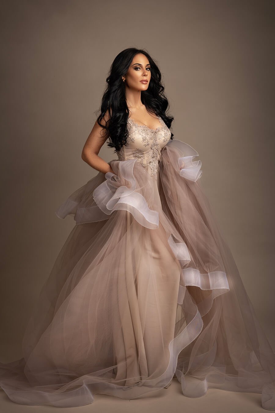 brunette model poses in a studio during a glamour photoshoot. she has a long tulle dress with a lot of bridal lace details on the top. 