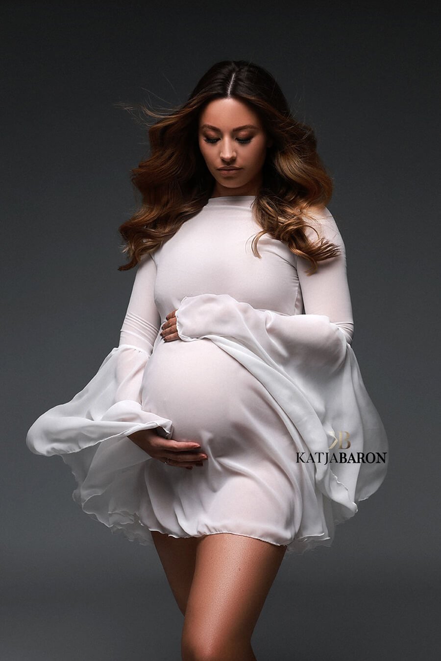 A pregnant woman is wearing a short wavy white dress. She has her hands around her belly and is looking down