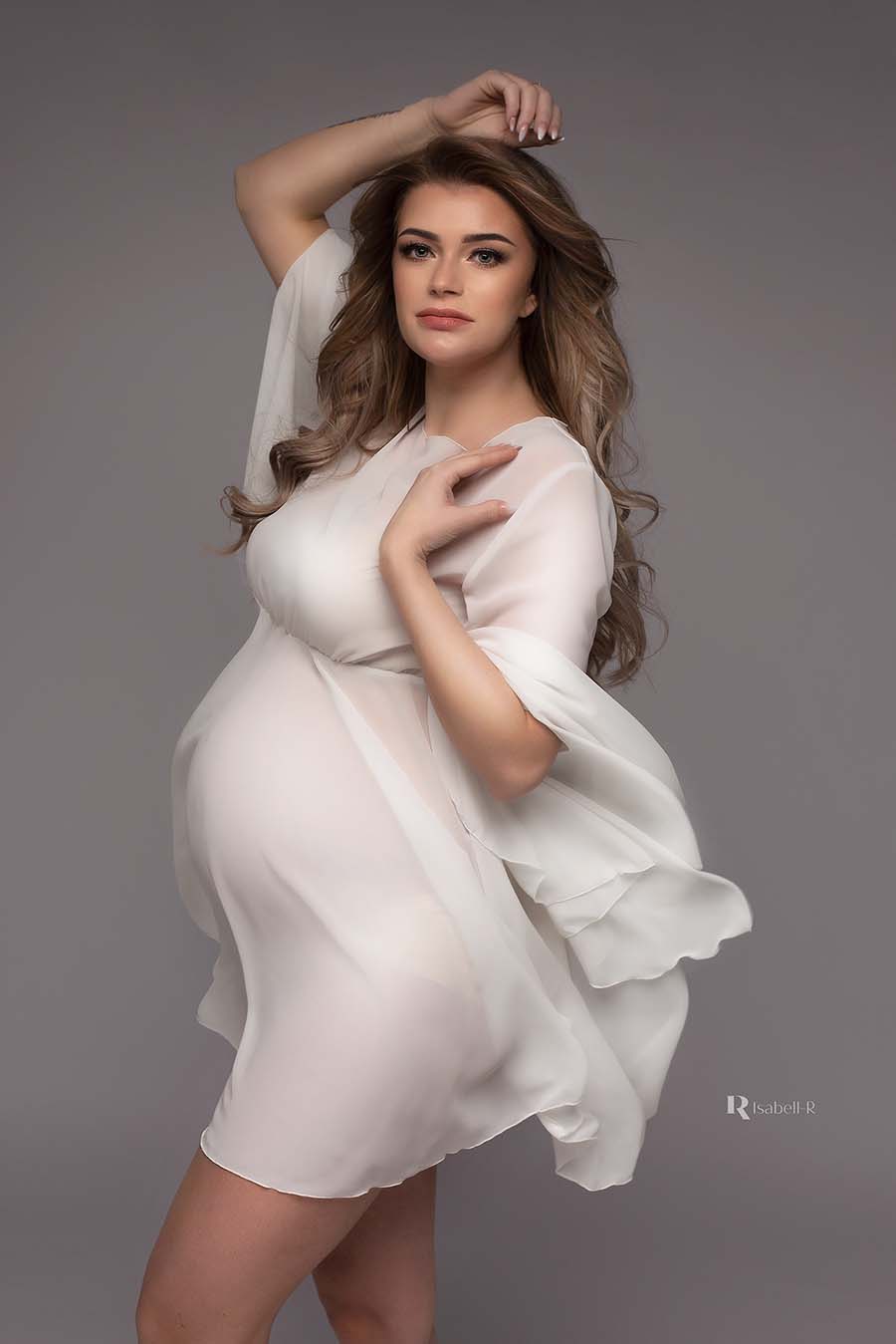 A pregnant woman is posing in a short white dress. The dress is made out of chiffon and is a little bit sheer. She has dark blonde hair.  The woman is posing with one hand above her head and the other one is laying by her shoulder. The dress is a light fabric. 