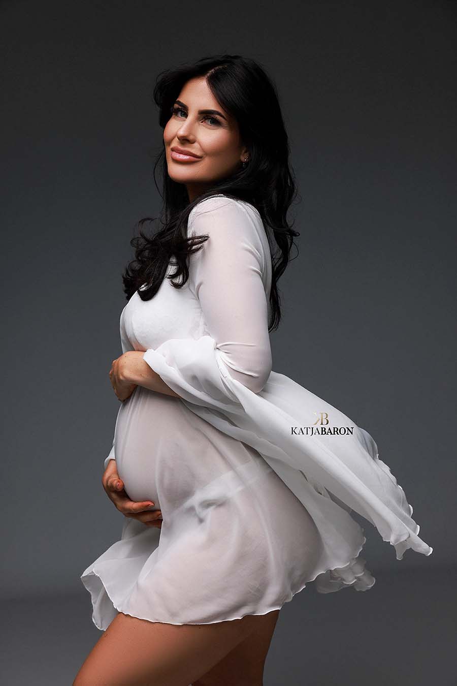 A model with  long black hair is posing for the camera. She is wearing a short white dress. The dress has a light chiffon fabric and is waving in the wind. She is pregnant and is posing with her hands around her belly. 