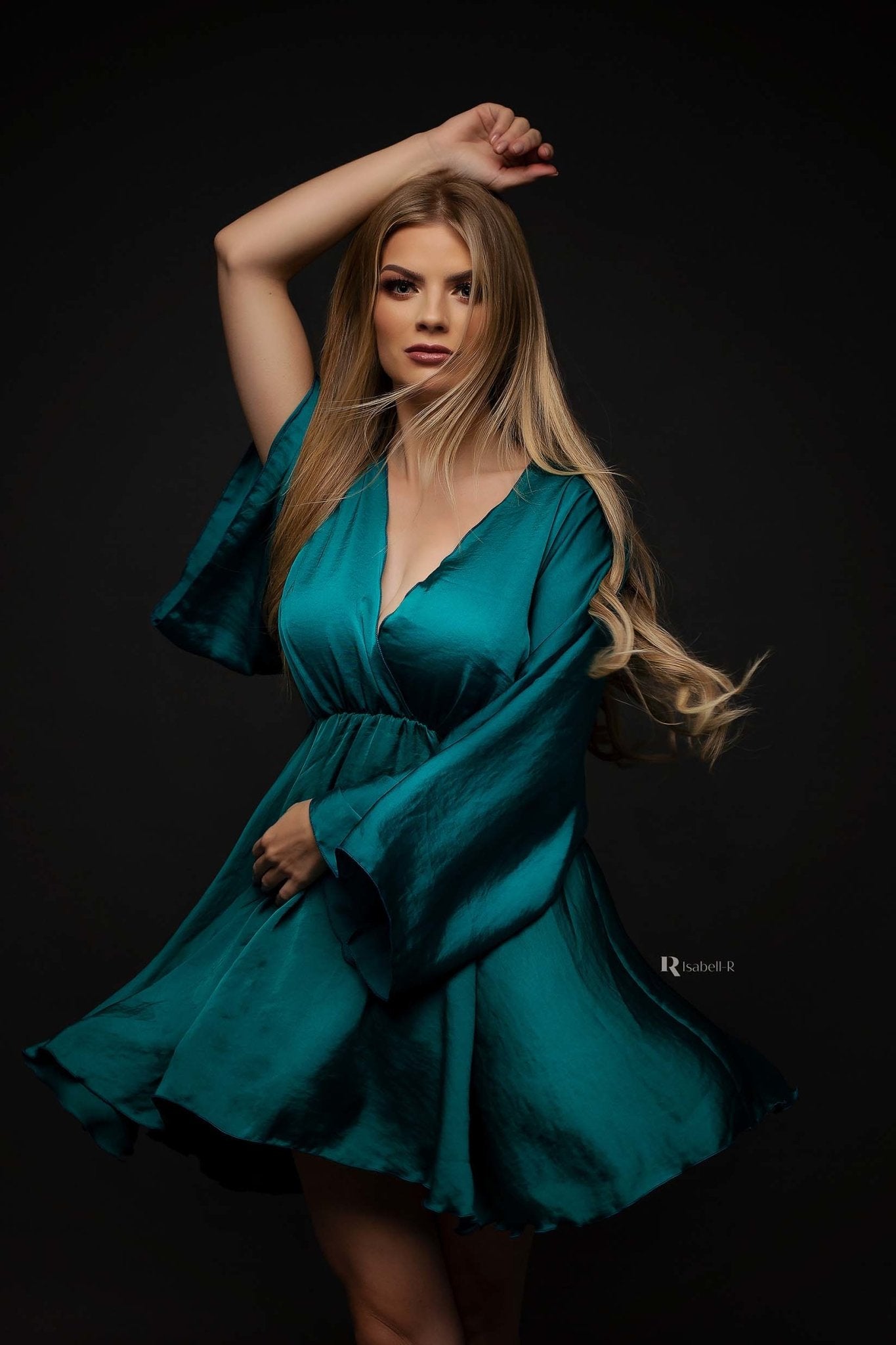 blond model poses wearing a short silky dress in emerald color. the dress has kaftan style sleeves and a low v cut neckline.
