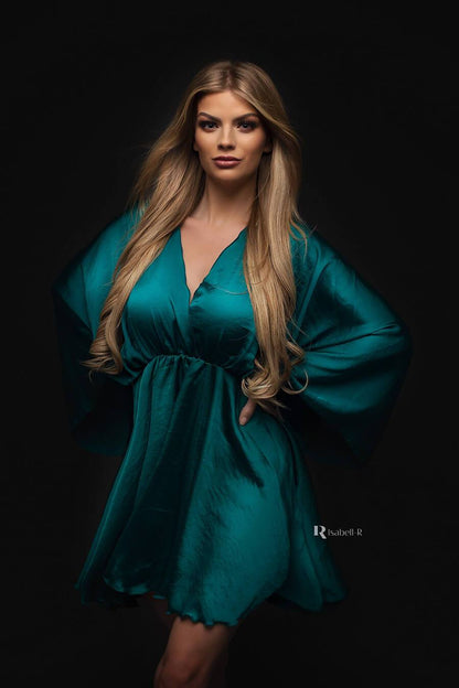 blond model poses wearing a short silky dress in emerald color. the dress has kaftan style sleeves and a low v cut neckline.