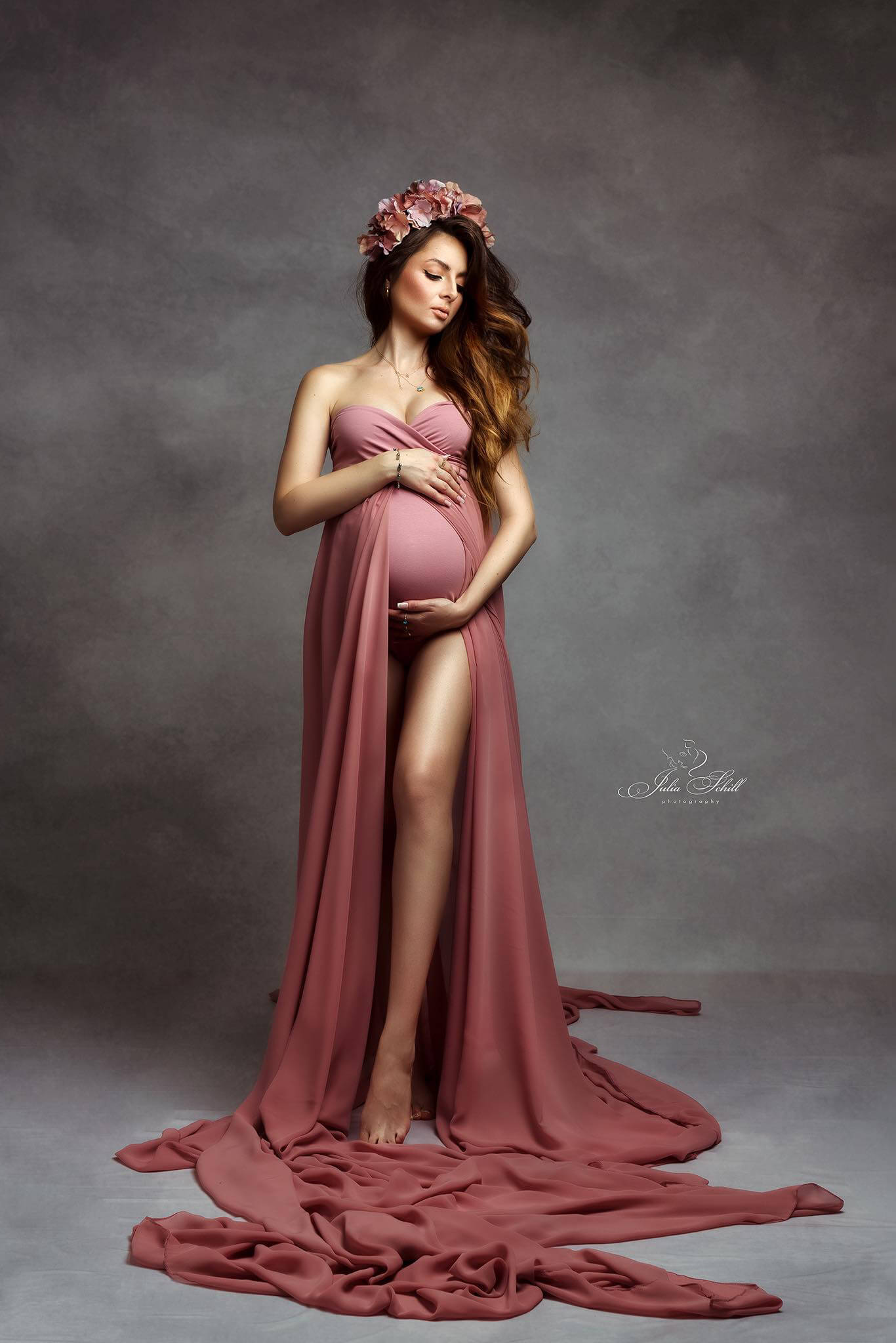 dark blond pregnant model poses in a studio against a grey background. the model wears a pink strapless maternity bodysuit. the bodysuit has a chiffon skirt attached. the skirt has a middle front split where one of her legs can be seen through. she has her eyes closed and holds her bump with both hands. 