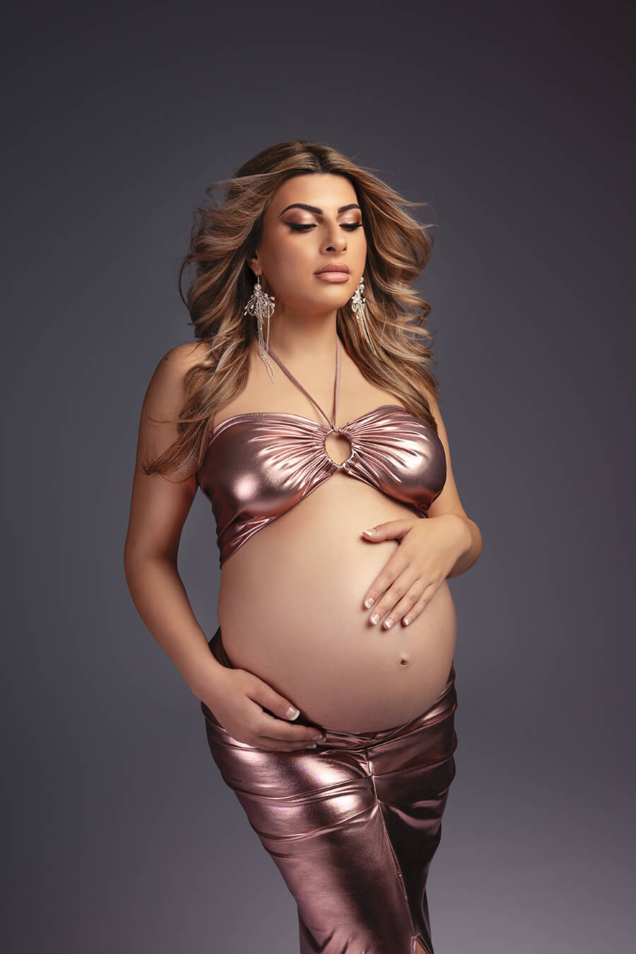 close up of blond pregnant model posing in a studio holding gently her belly bump. she is wearing a lamee set in dusty pink color. the top has a keyhole peekaboo detail and the skirt is tight