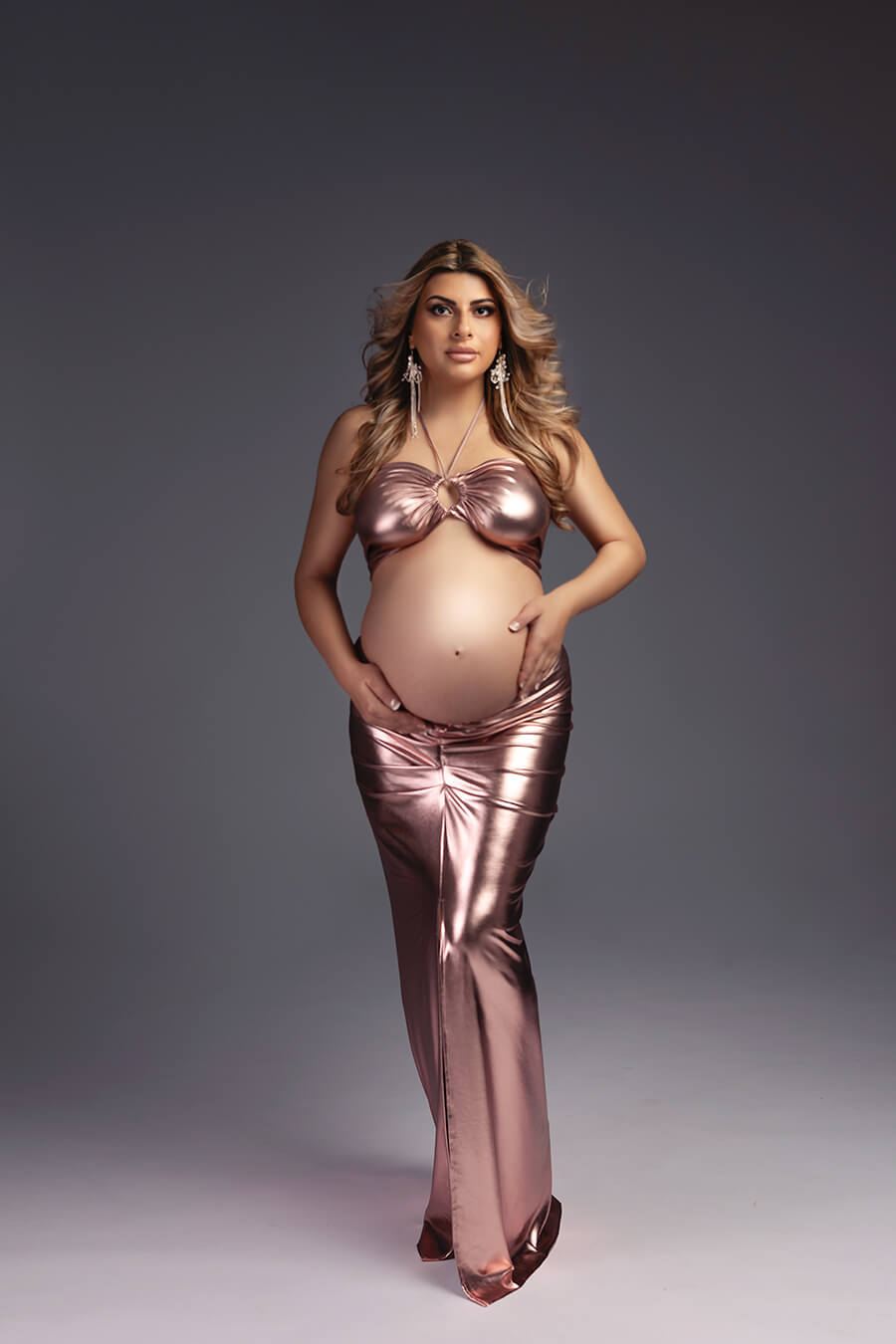 blond pregnant model poses in a studio holding gently her belly bump. she is wearing a lamee set in dusty pink color. the top has a keyhole peekaboo detail and the skirt is tight
