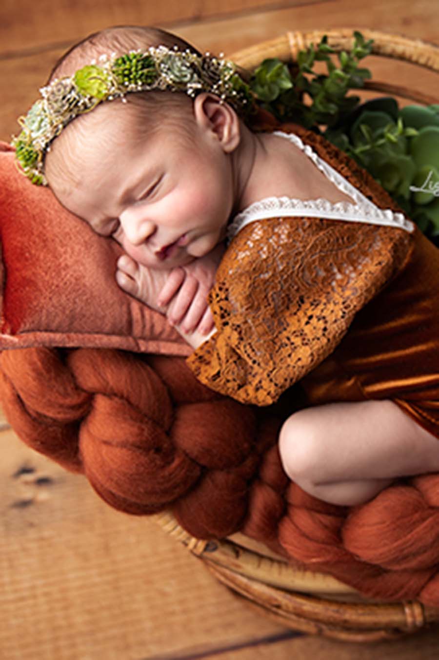 A baby is laying on a blanket in a little basket. The baby is wearing a hairband with fake green plants and the linden romper cognac.