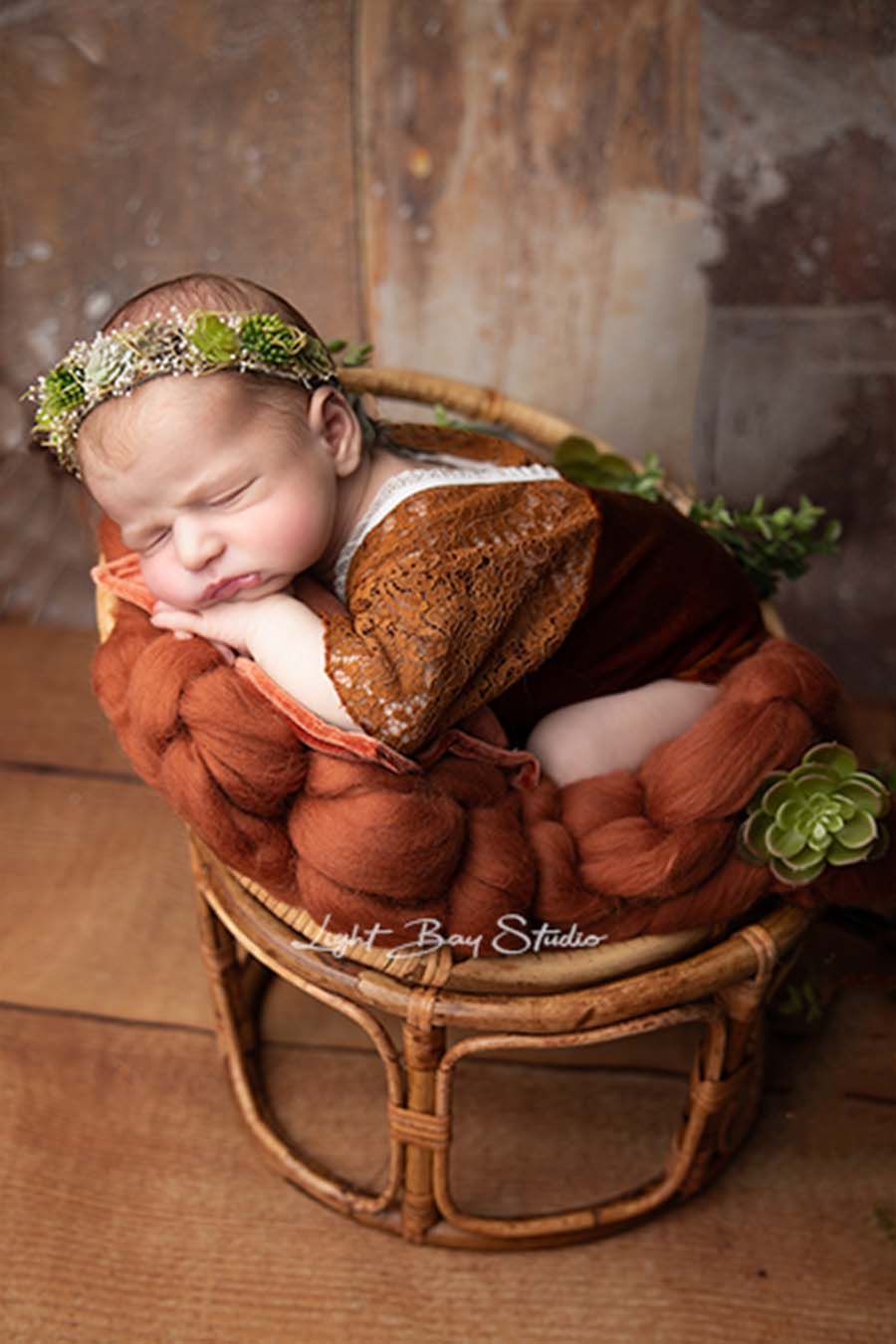 A baby is laying on a blanket in a little stool. There are fake plants. The baby has his eyes closed and is wearing the linden romper cognac. 