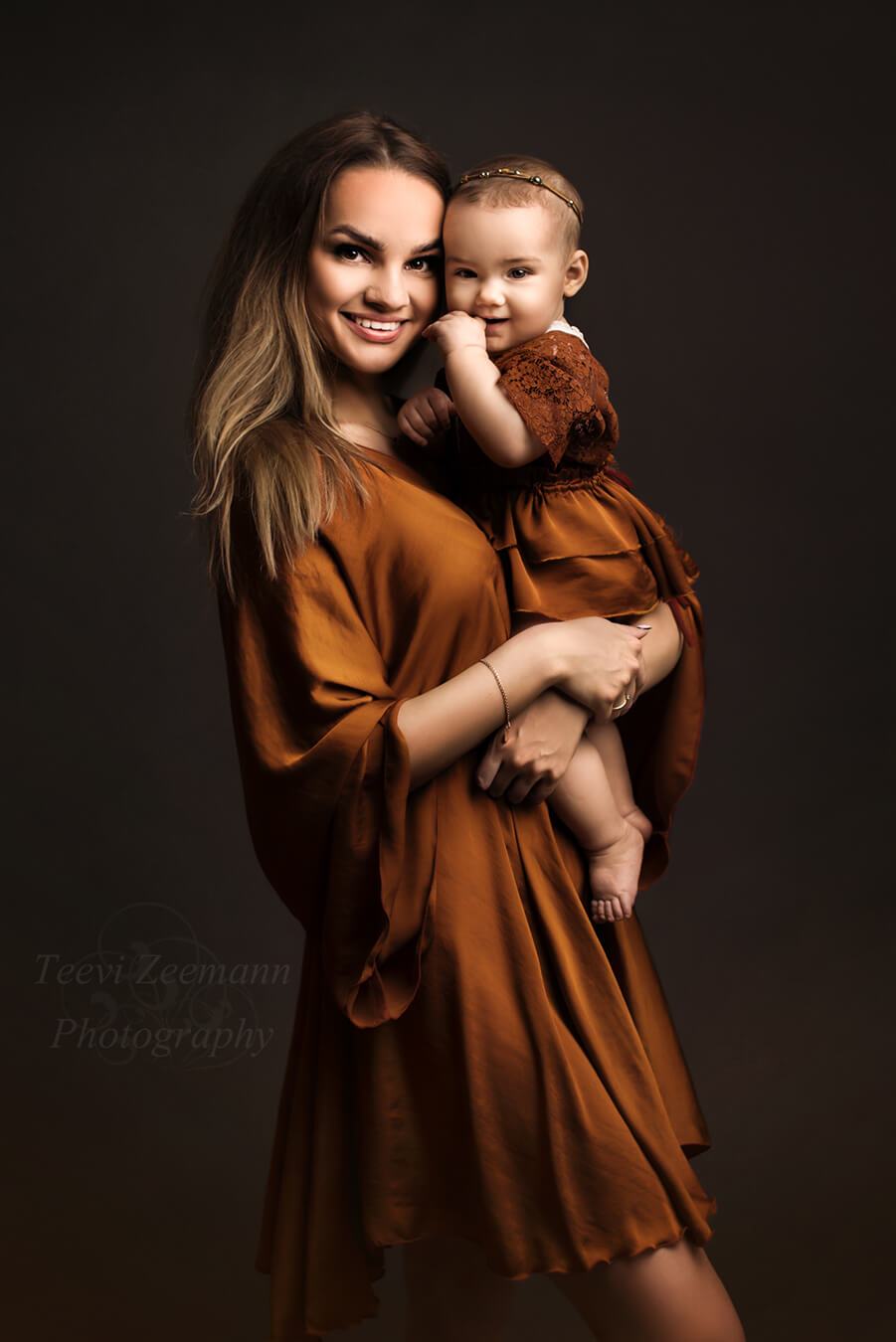 A mother is holding her baby girl. They are wearing matching outfits in the color cognac. The mother is wearing the lilibet dress and the baby is wearing a romper with a skirt on top of i. the skirt and the dress are both in silky material and the romper is lace.