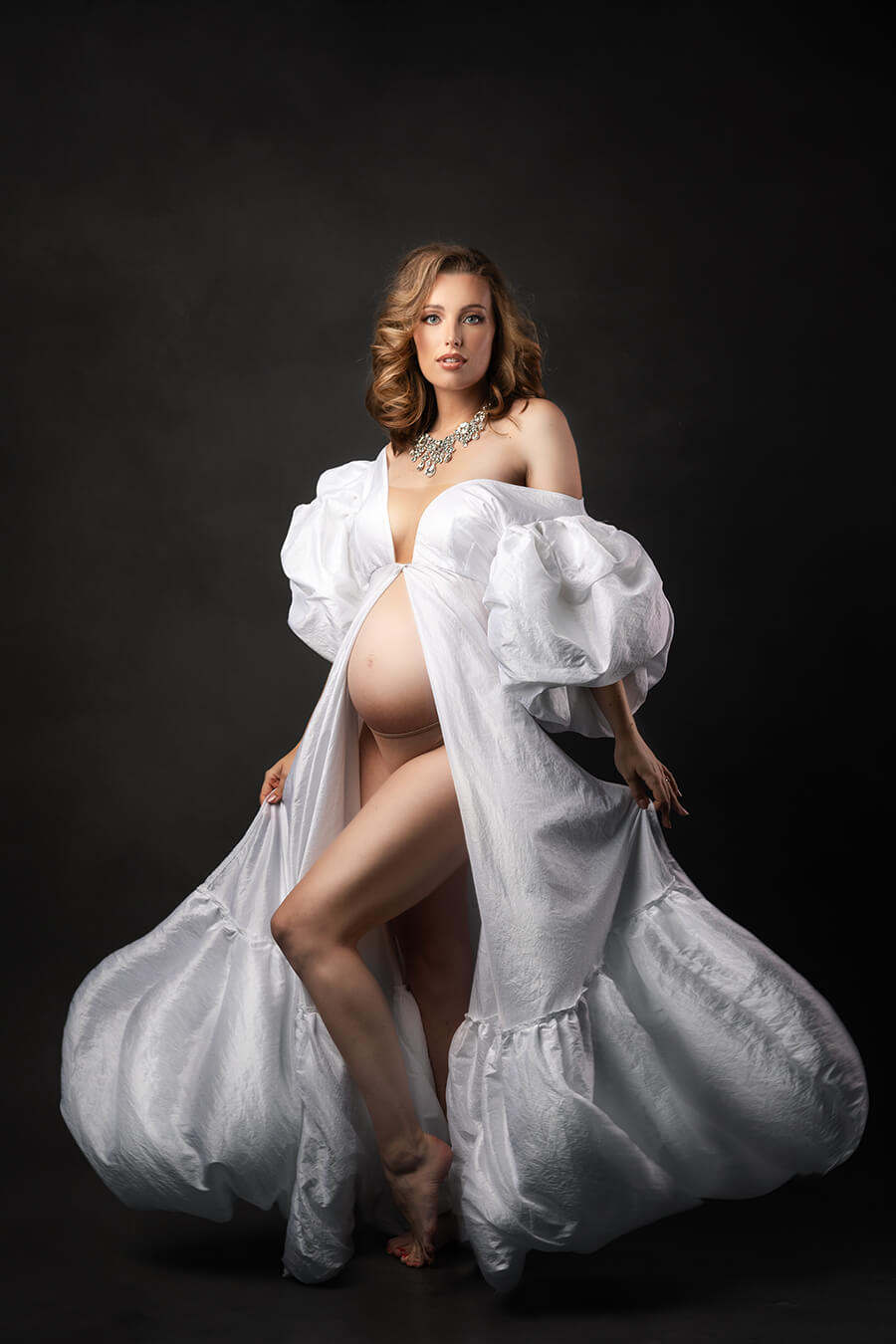 pregnant model in a studio poses wearing a long and open in the middle white dress made of taft. the dress has exaggerated sleeves