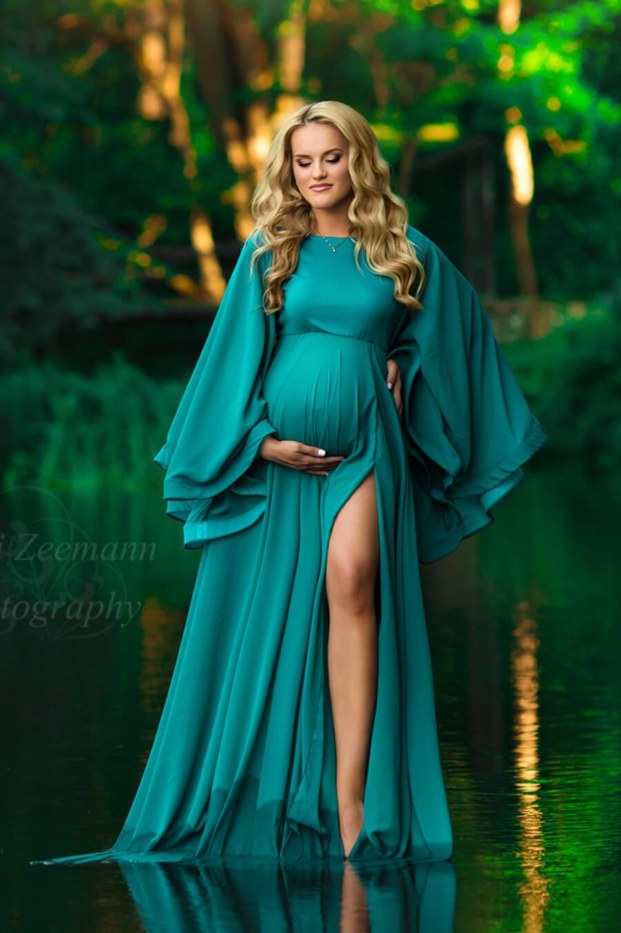 a pregnant woman with blonde hair is standing outside in a small lake. The trees are green behind her. She is wearing a maternity dress with wide sleeves. The skirt is open by her leg 