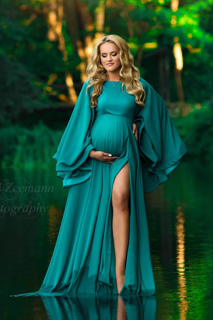 a pregnant woman with blonde hair is standing outside in a small lake. The trees are green behind her. She is wearing a maternity dress with wide sleeves. The skirt is open by her leg 