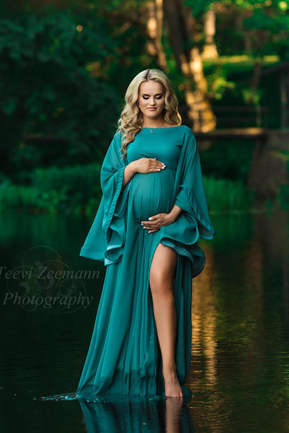 A woman is standing in a lake. She is wearing a long dress in the colour petrol. She has long blonde hair and looking down. The dress has a split by her left leg 