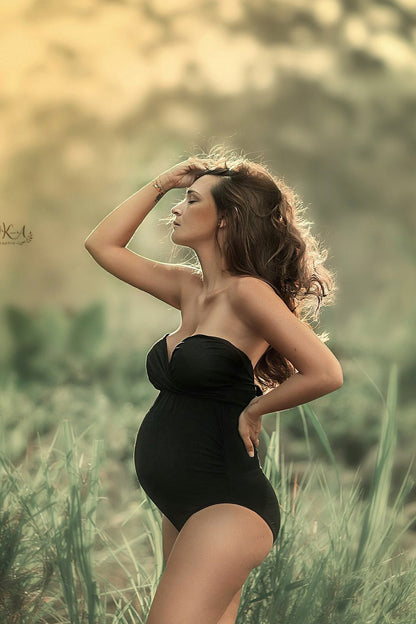 This model is faced to the side. She is standing outside in tall grass. She is wearing a black bodysuit with a bandeau top. She has brown curly hair
