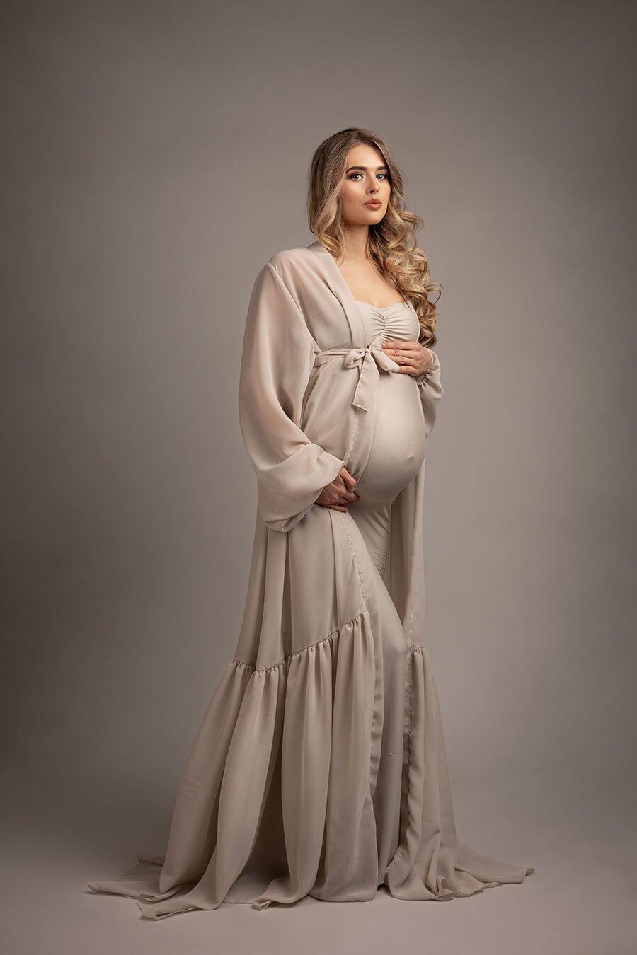 pregnant blond model poses in a studio holding her baby bump and looking to the camera. she is wearing a long chiffon robe in sand color and a matching long tight dress underneath. 