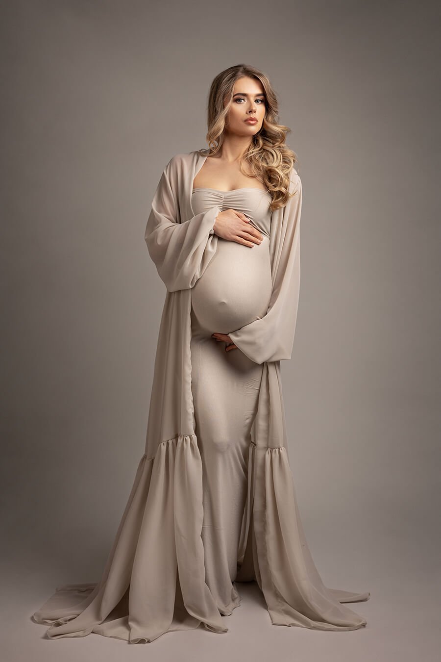 pregnant blond model poses in a studio holding her baby bump and looking to the camera. she is wearing a long chiffon robe in sand color and a matching long tight dress underneath.