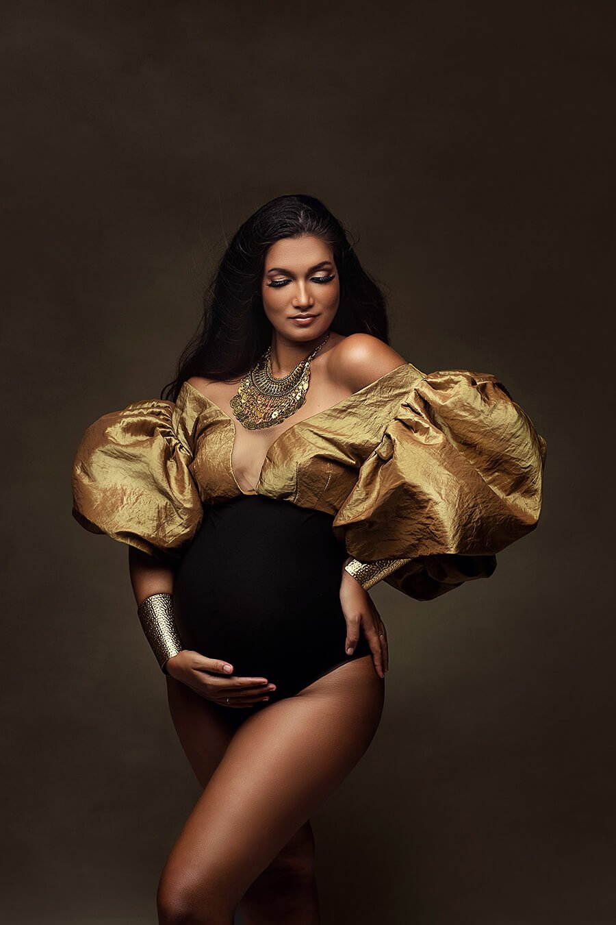 dark haired pregnant model poses in a studio wearing a bodysuit in two colors. the top is made of golden taft and the bodice of black jersey. the top has exaggerated puff sleeves.