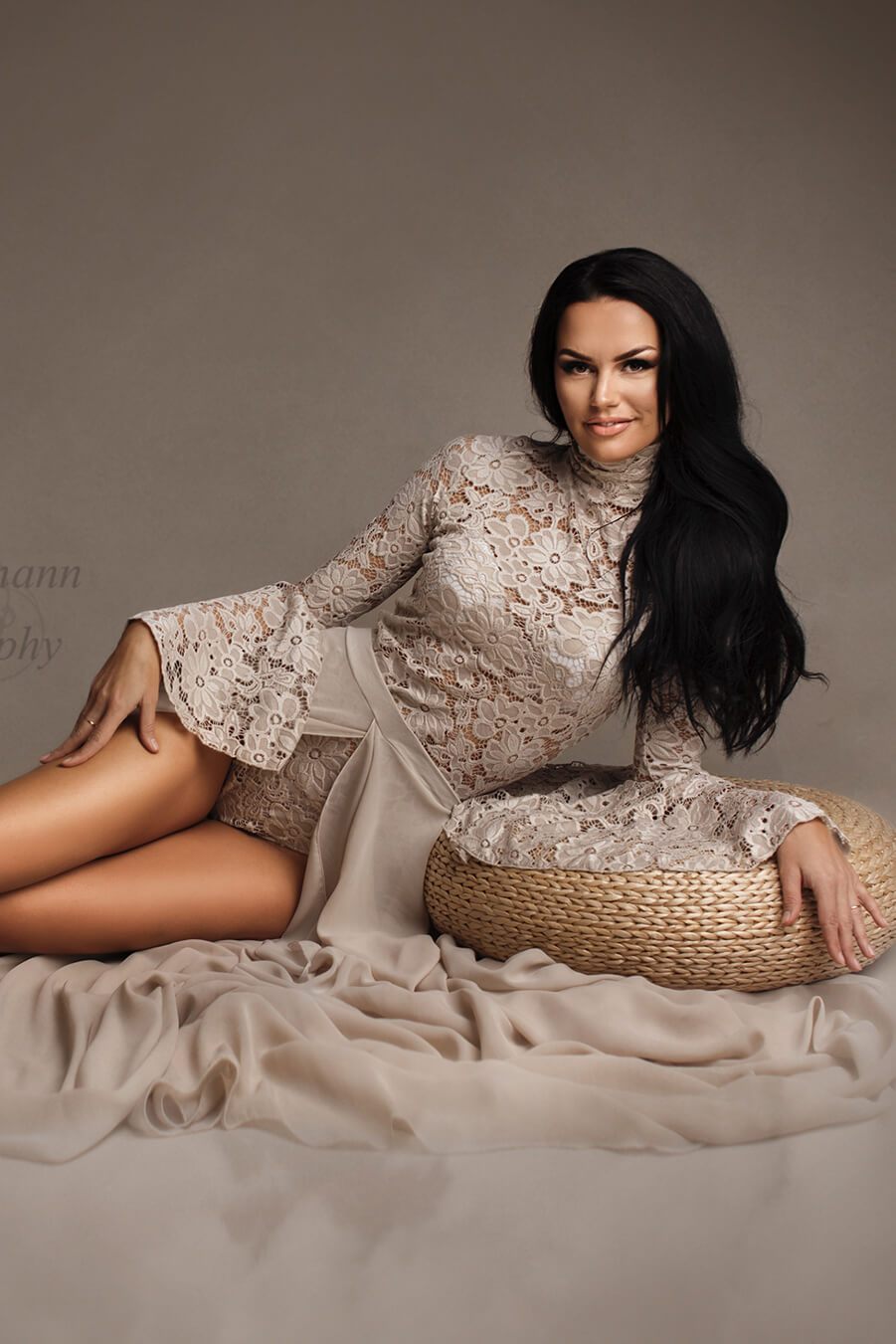 A woman with black hair is laying on the ground. She is leaning on a small box. She is wearing a bodysuit with a skirt in the colour sand