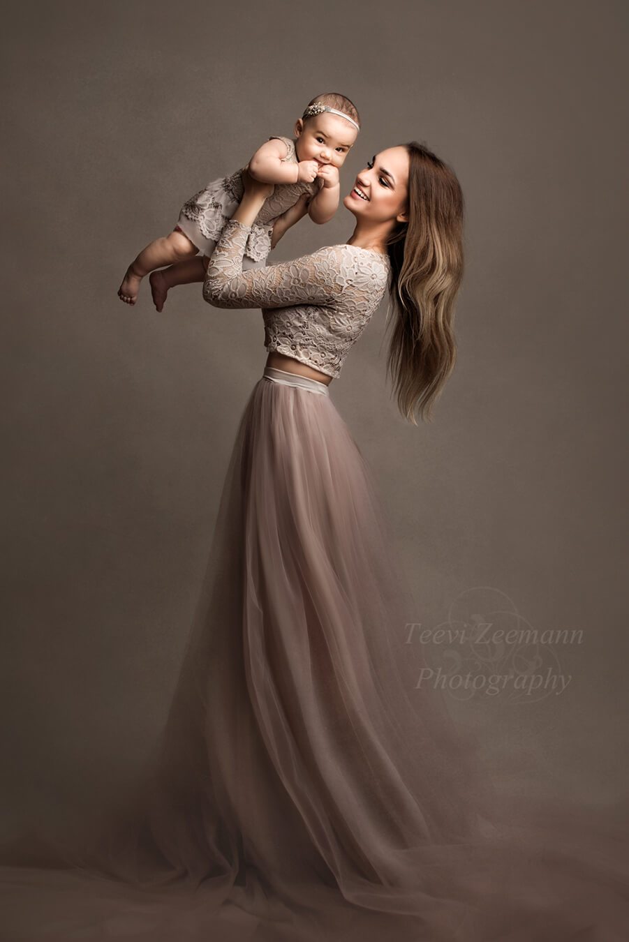 Dark blond model poses with her baby during a photoshoot. She wears a lace top with long sleeves and a tulle long skirt. Her baby has a matching dress also made of lace. She holds her baby up while smiling at her.