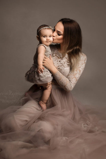 Dark blond model poses with her baby in a studio photoshoot. She holds her baby while kissing her cheek. The mother wears a long sleeved lace top and a matching tulle skirt while the baby wears a little dress also in lace.
