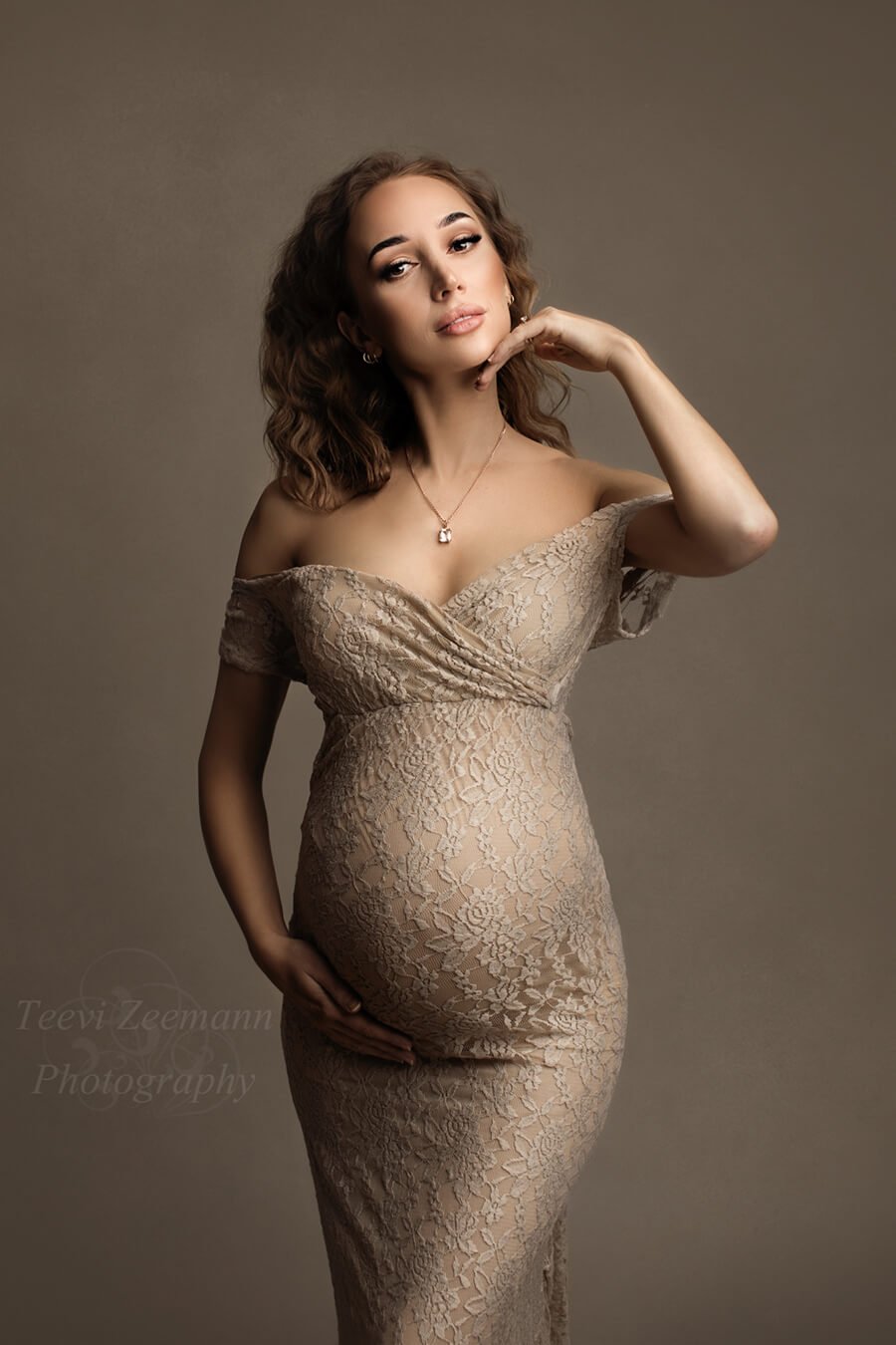 pregnant model faces the camera and touches her chin with one hand while the other is resting on her bump. she is wearing and off shoulder, short sleeves sweetheart neckline dress in sand color and made of lace. 