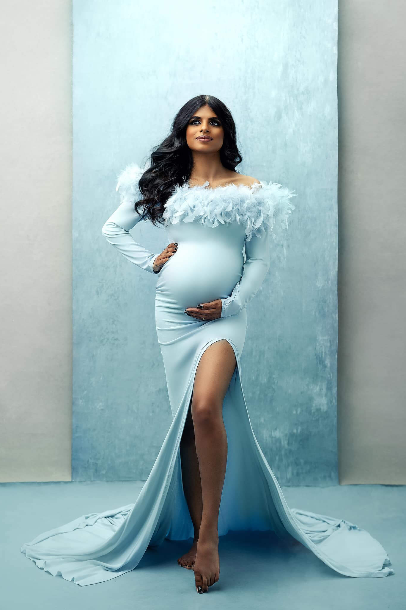 A pregnant woman is posing in in long light blue dress. The dress is off shoulder with a split by the leg. The dress is made from a jersey fabric and has a tight fit. The top of the dress has feathers for a chique look.