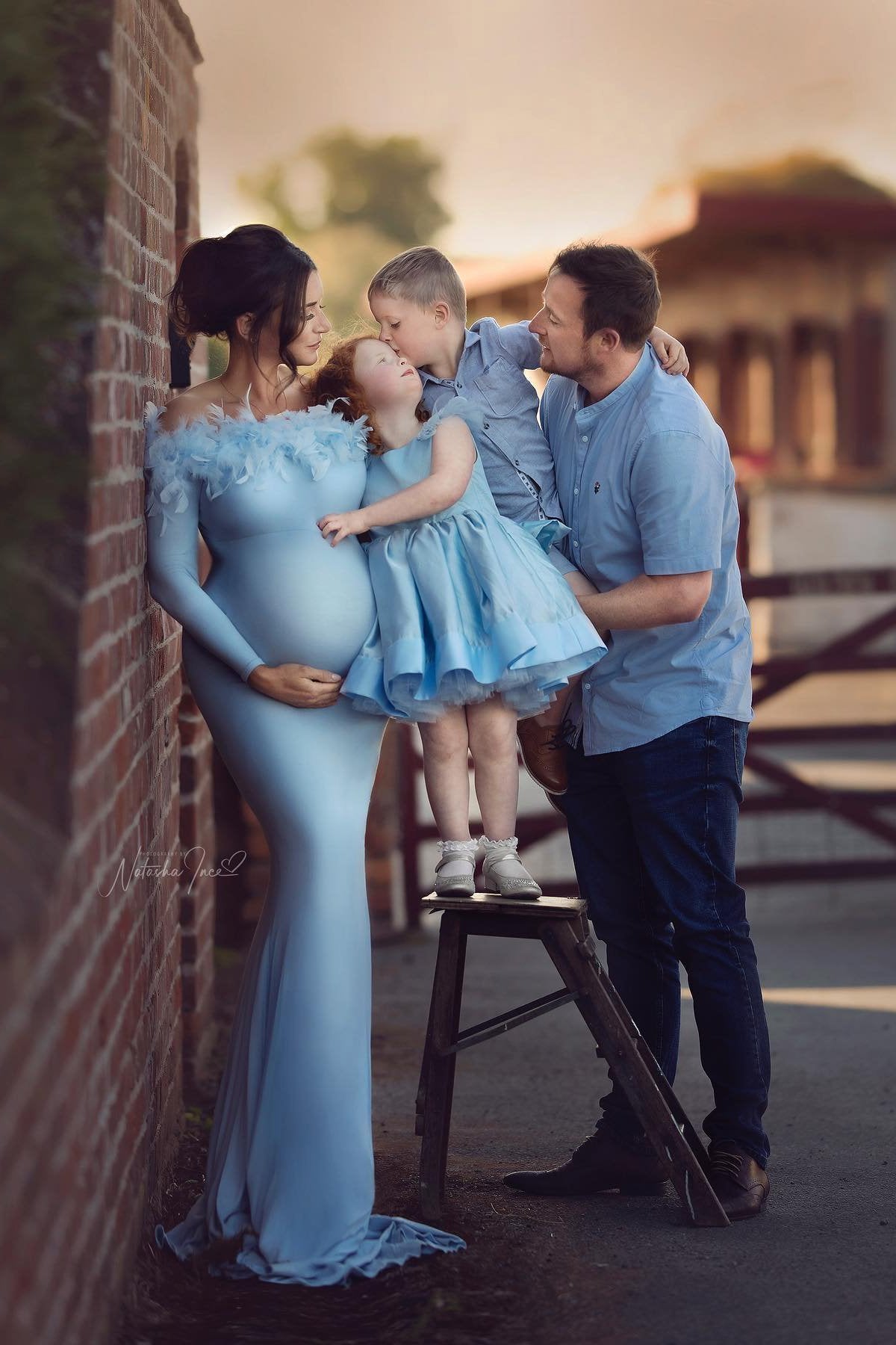 This is a family photo taken outside by a ranch. There is a mother, a dad and their two children. One boy and one girl. They are all wearing matching light blue outfits. Our product is the dress that the mother is wearing. The dress is off shoulder and a tight fit. The dress has feathers at the shoulder band. 