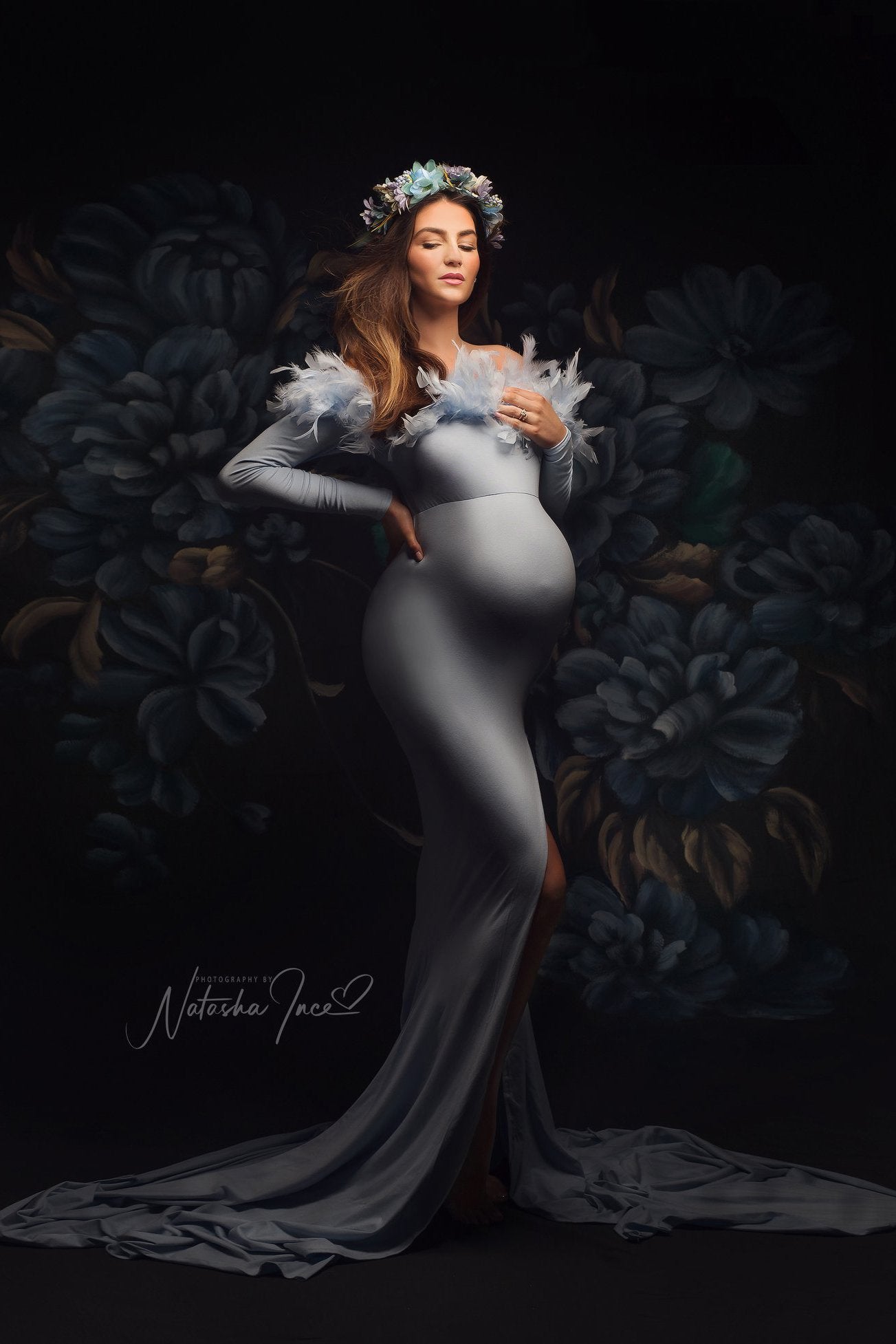 A pregnant model is posing in a studio. There painted blue flowers in the background. The woman is wearing a long blue dress. the dress is a off shoulder model and the top part by the shoulder has a line with feathers. She is wearing a hairband with matching blue flowers. 