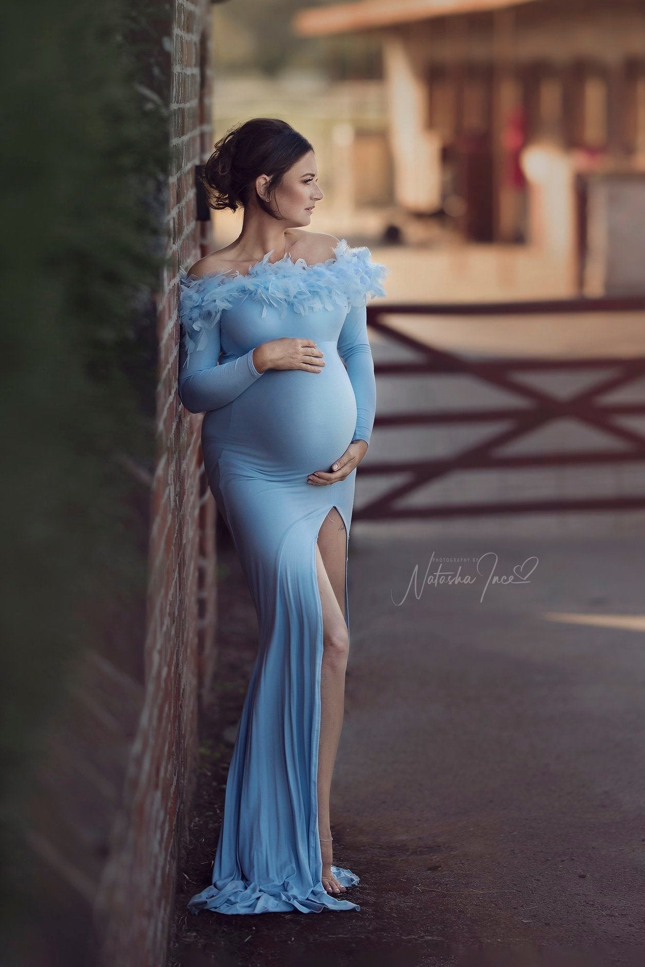 A pregnant woman is standing outside a ranch. She is leaning against a brick wall. She is wearing a long dress in the color light blue. The dress is off shoulder and has a split. The sleeves are long and the top part is covered with feathers. 