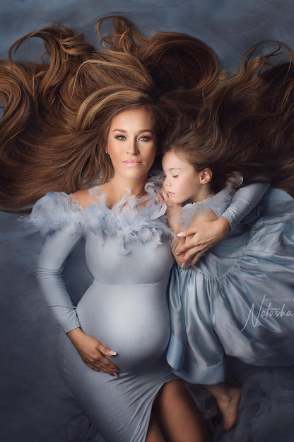 A pregnant mother is laying with her daughter on the ground. They are hugging each other. They are both wearing a dress in a matching light blue color. The dress that the mother is wearing has a tight fit and is off shoulder. The top has a band with feathers. The girls dress has a princess look. 