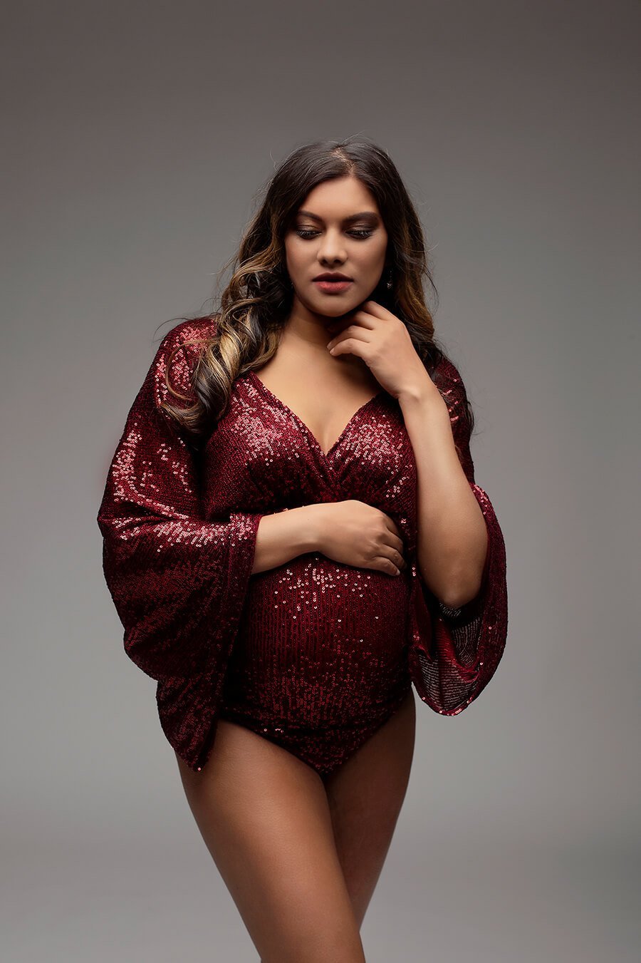 This pregnant woman is wearing a sparkle bodysuit. She has one hand on top of her belly and one by her neck. She is looking down at the ground. 