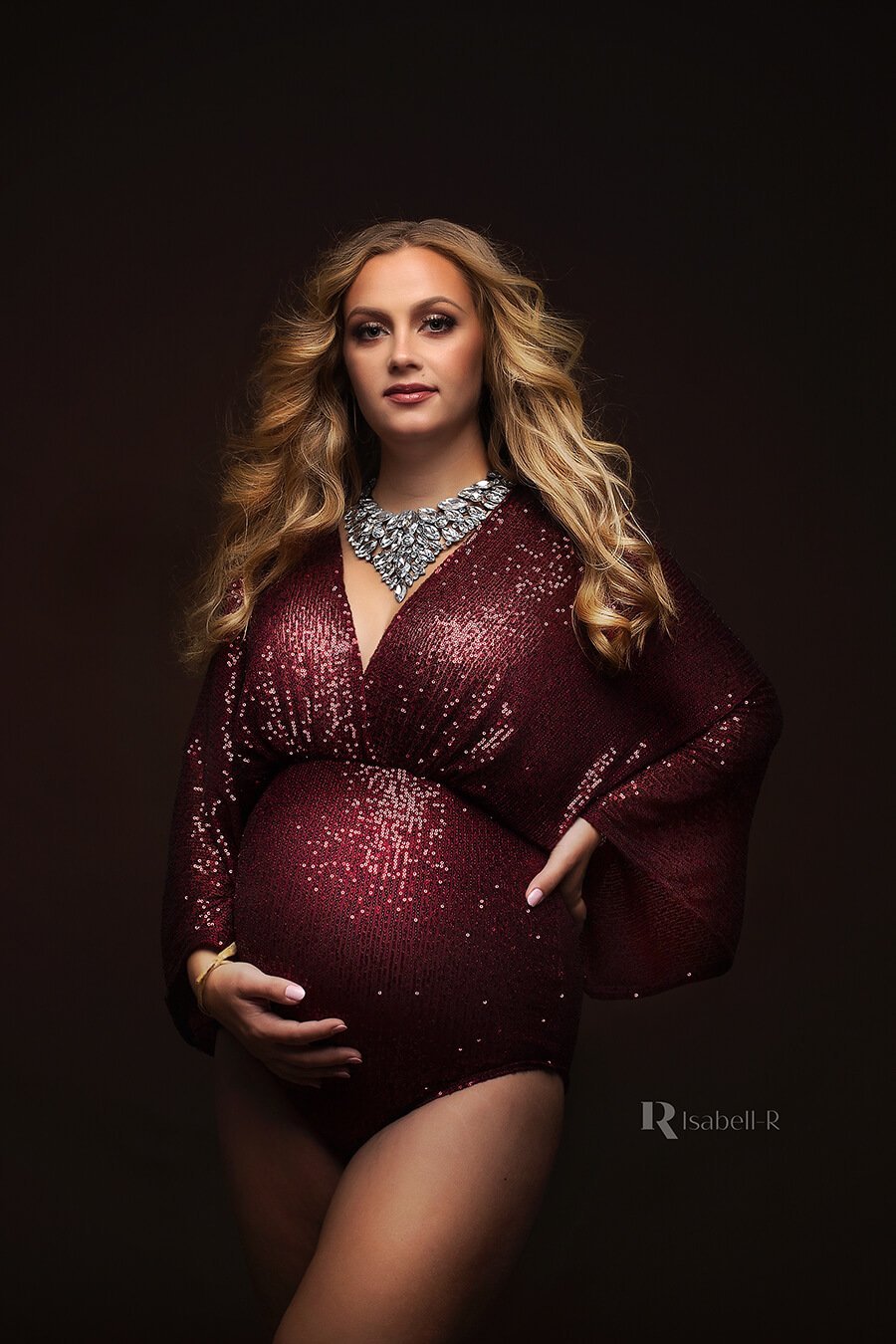 A maternity model wearing a sparkle bodysuit in the colour bordeaux. The bodysuit is boho styled and she is wearing a big necklace with it. She has long blonde hair