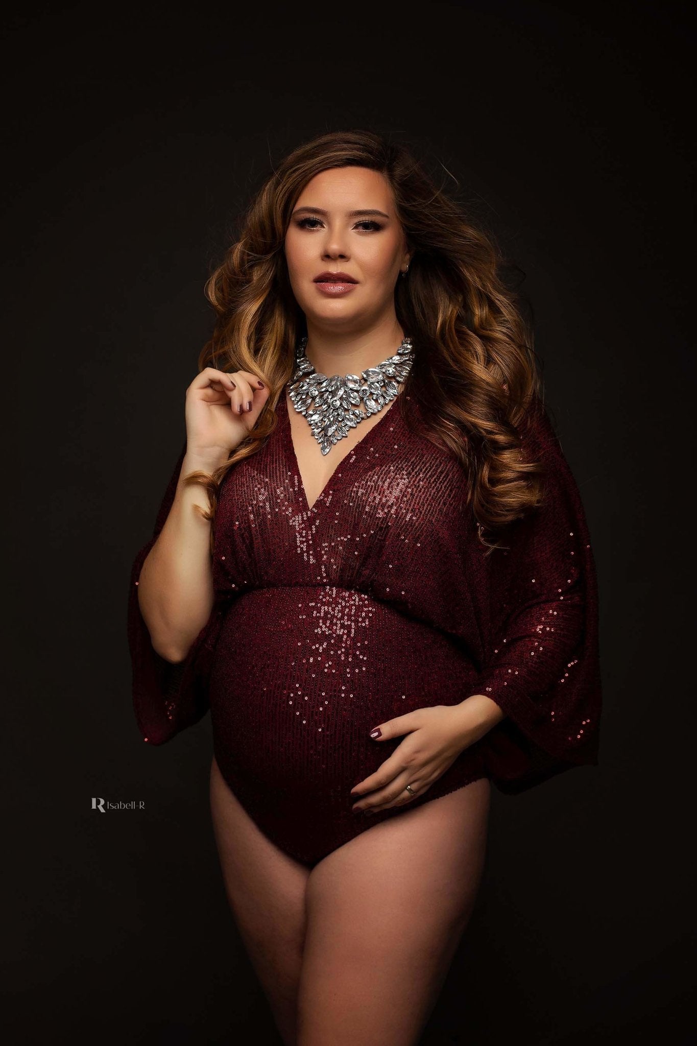 A woman with long brown hair is wearing a bordeaux sparkle bodysuit. The bodysuit is boho styled. She has one hand by her belly and one in her hair. Her nails are painted in the colour red