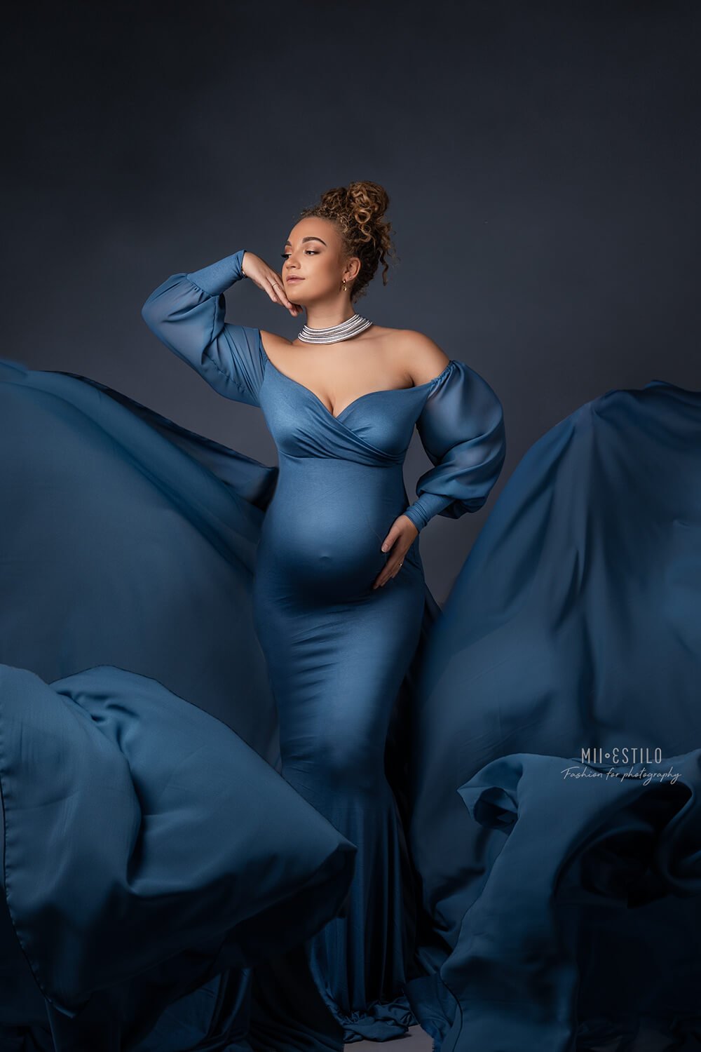 pregnant model poses in a studio wearing a long denim dress with an adjustable sweetheart top and bishop sleeves. 