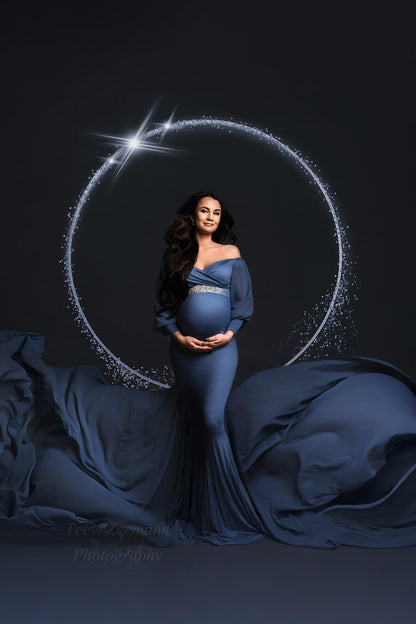 brunette model poses in a studio wearing a denim maternity dress with bishop sleeves and an adjustable sweetheart top. the dress has a long chiffon train. the model poses in a studio and wears a sash to match the style. 