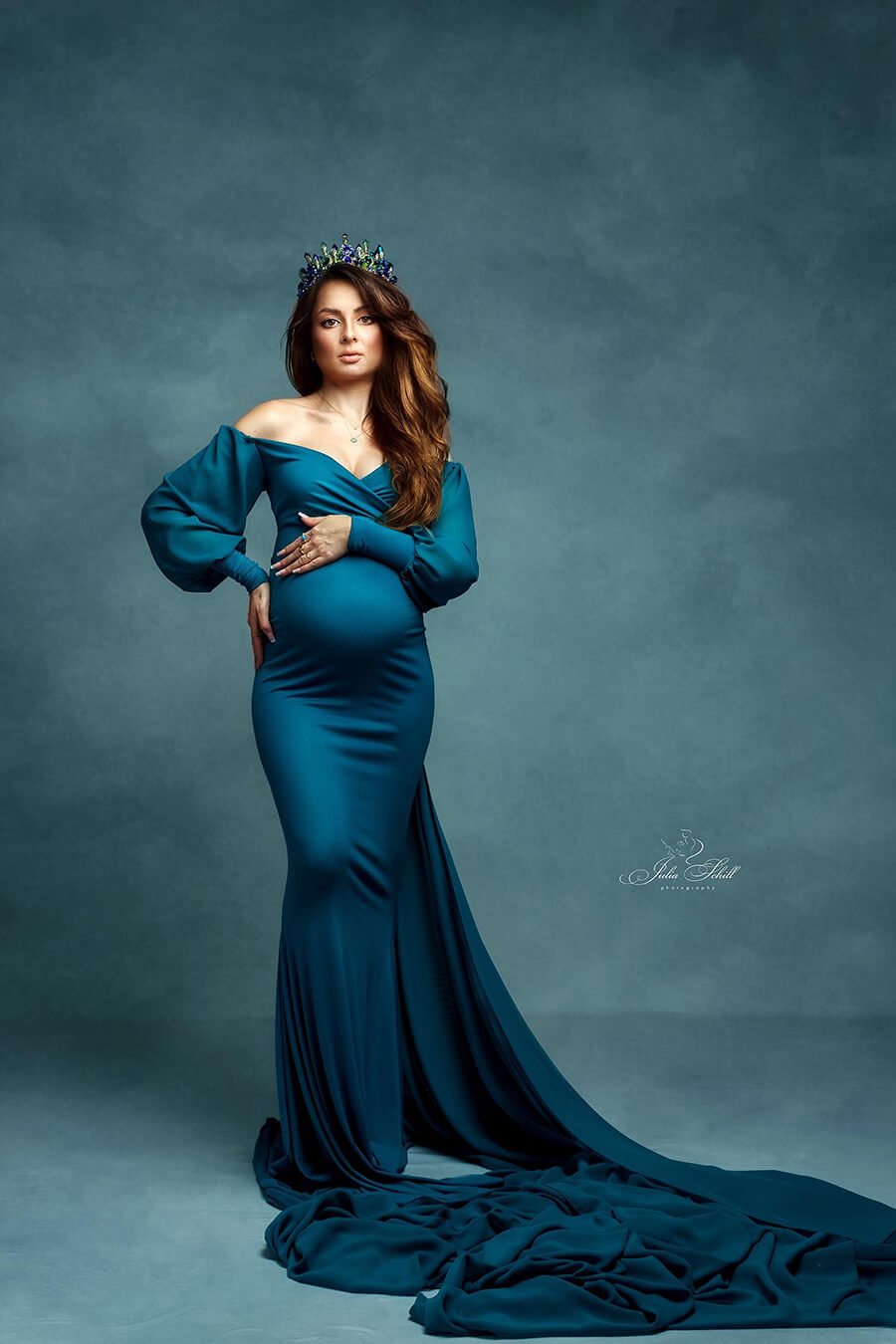 dark blond pregnant woman with long hair poses wearing a petrol colored dress. the dress features a very long chiffon train to create several effects while being photographed. the model looks straight to the camera while holding her baby bump with both hands.