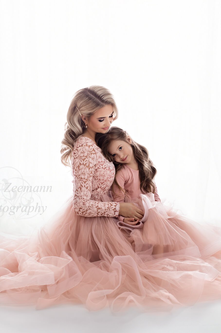 A mother is sitting on the ground wearing a pink tulle skirt. Her little daughter is sitting on her lap. She is also wearing a pink tulle dress