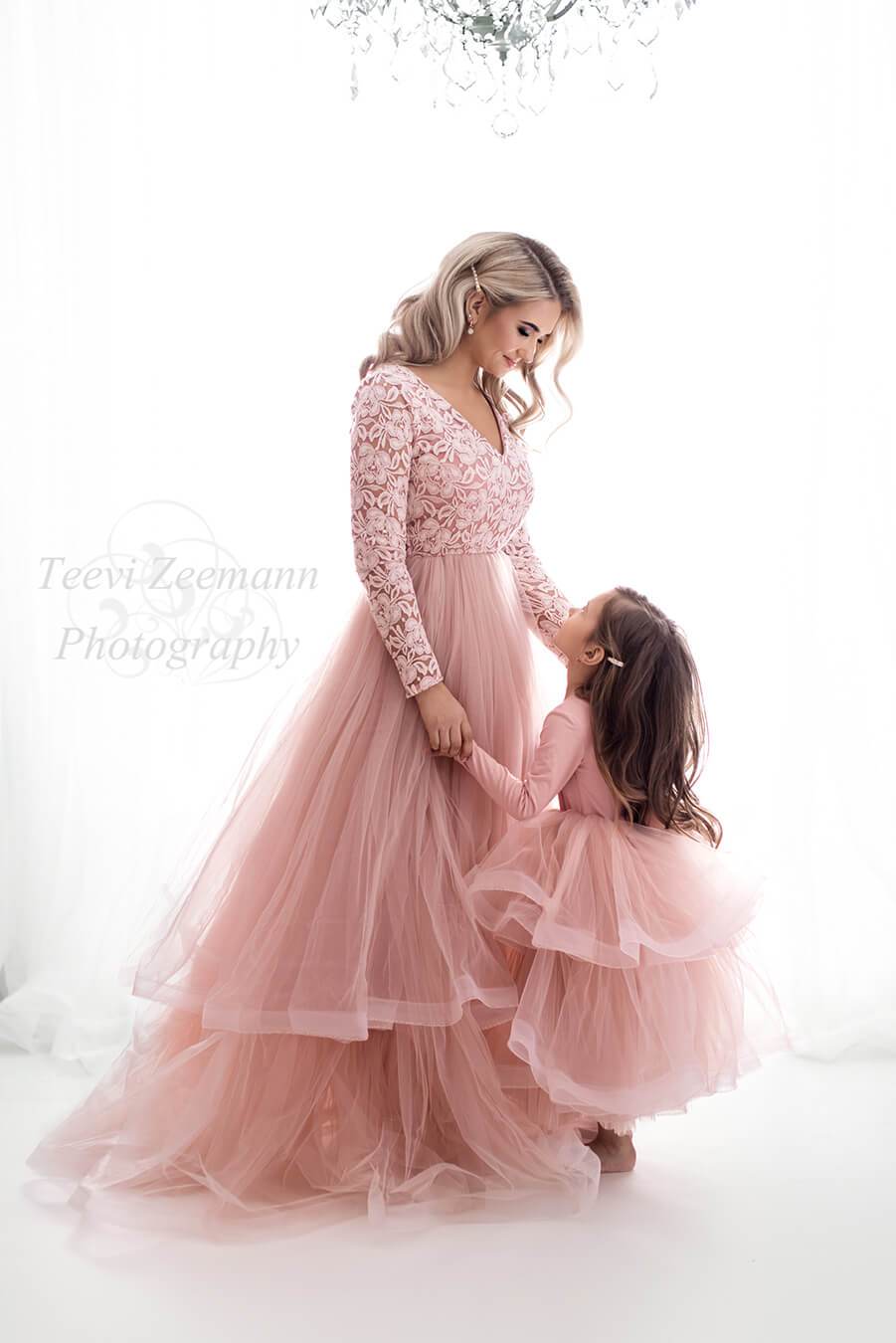 A mother is wearing a pink dress with a lace top and a tulle skirt. The little girl is looking at her mother and is wearing a dress with a big tulle skirt