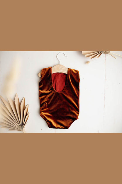 The back of a baby romper. The romper is laying on a white background with paper leaves. The romper is from velour and in the colour cognac