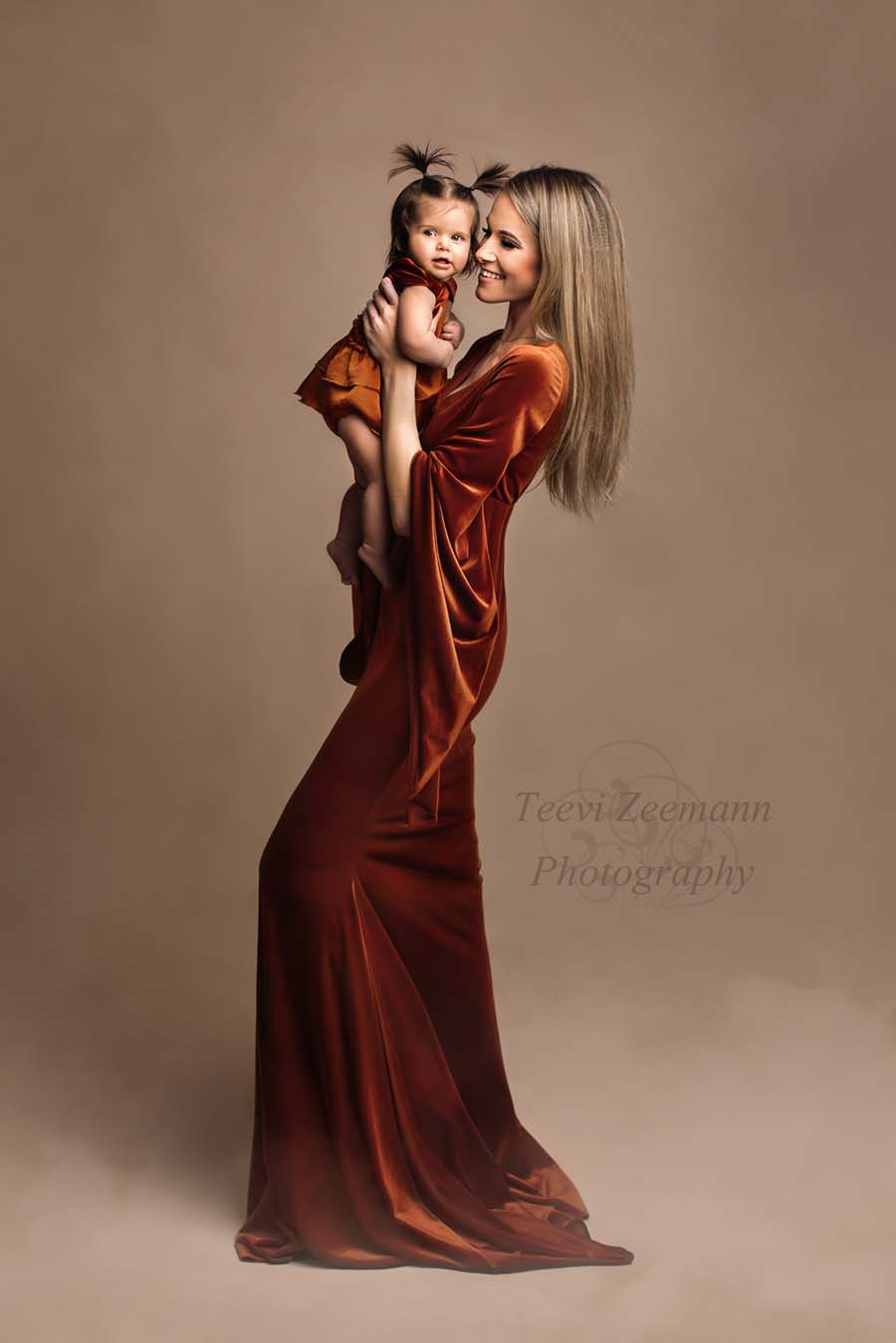 A mother is holding her little girl. They are both wearing a velour outfit in the colour cognac. The girl is wearing a little romper and the mother a long dress