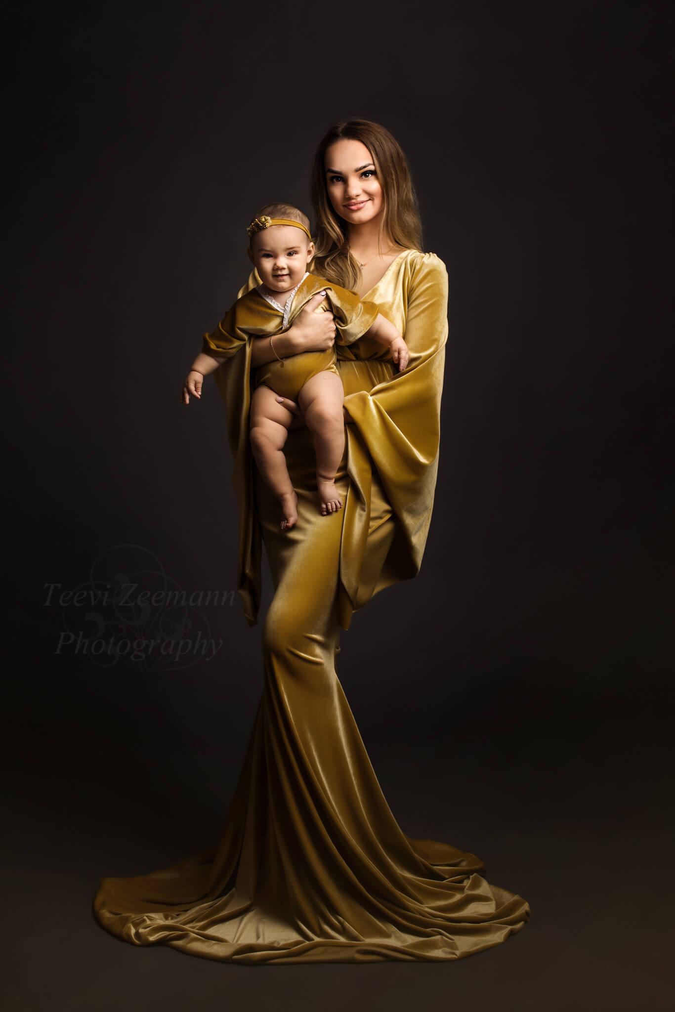A gorgeous mother is holding her baby. They are both wearing a velour outfit in the color amber. The mother wears a long dress with boho sleeves . The baby wears a romper and has a little headband on 