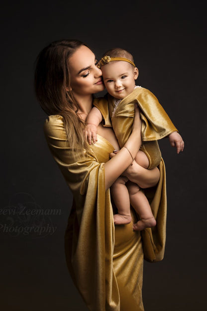 A woman is holding her baby. She is looking down at him with a little smile. The baby is looking towards the camera. They wear matching outfits in the color amber. The baby wears a little romper and the mother an long dress