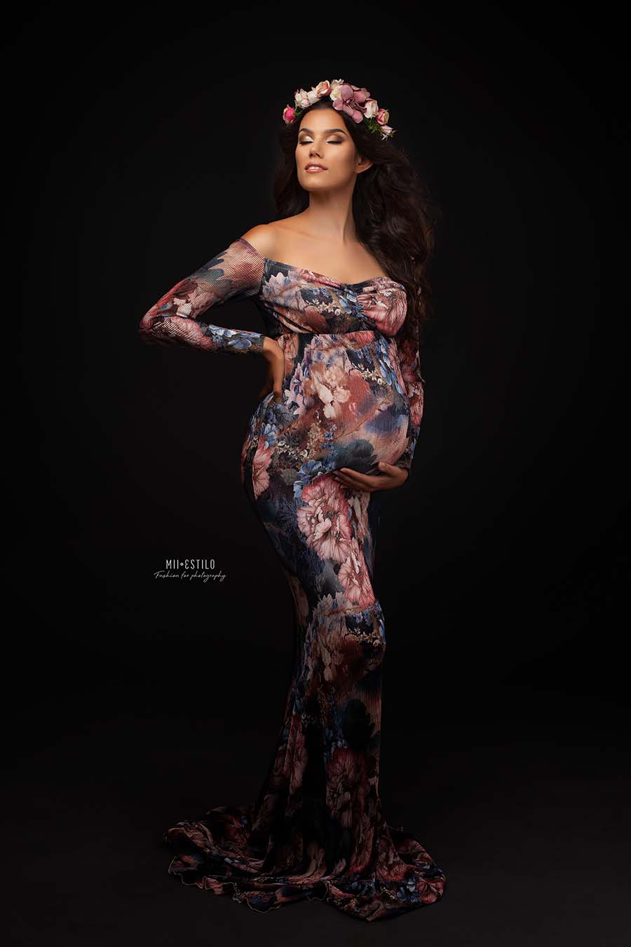 brunette model poses during her maternity photo shoot in a studio wearing a long dress with long sleeves, off shoulder top and sweetheart neckline and a matching flower crown. the dress has a flower pattern and it is very tight. the model has her eyes closed and faces up. 