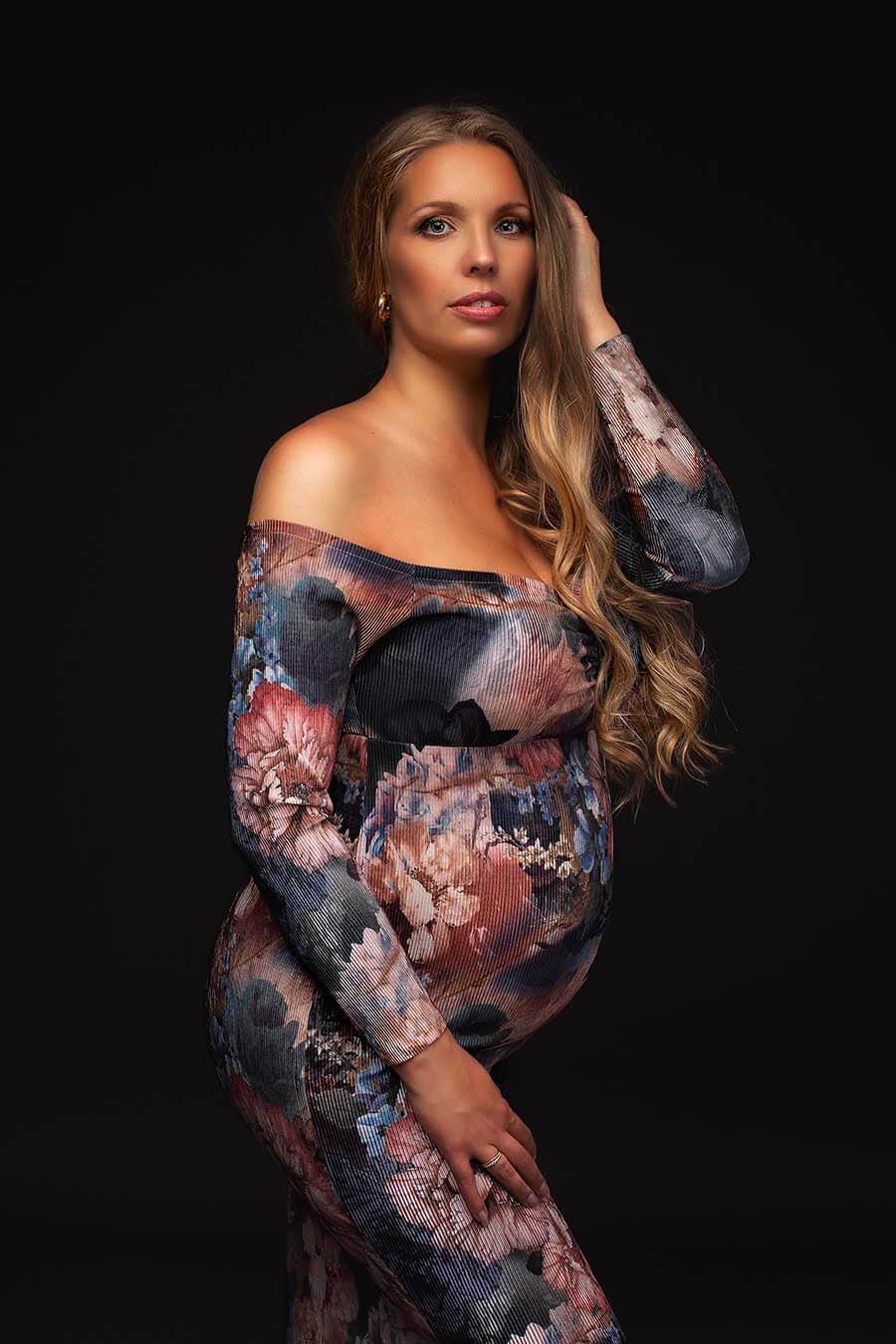 blond model poses during her maternity photo shoot in a studio wearing a long dress with long sleeves, off shoulder top and sweetheart neckline. the dress has a flower pattern and it is very tight.