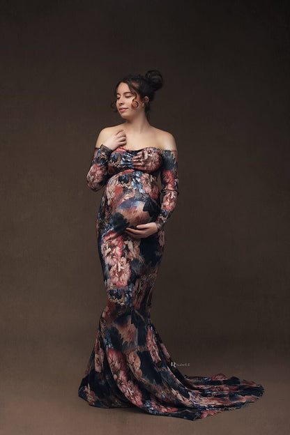 pregnant model poses in a studio wearing a long tight dress with flower pattern. she holds her bump with one hand and the other rests in her chest area. the dress has an off shoulder top, sweetheart neckline and long sleeves. 