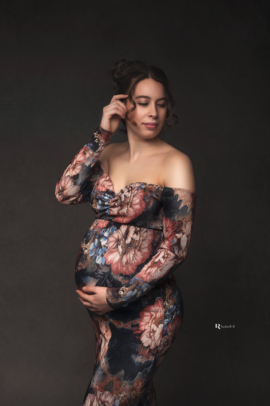 pregnant model poses in a studio wearing a long tight dress with flower pattern. she holds her bump with one hand and the other touches her hair. the dress has an off shoulder top, sweetheart neckline and long sleeves.