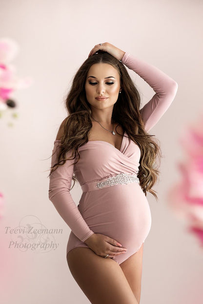 A pregnant model is wearing a pink bodysuit. She has one hand under her belly and one hand on her head. She has long brown hair and is wearing a little necklace around her neck 