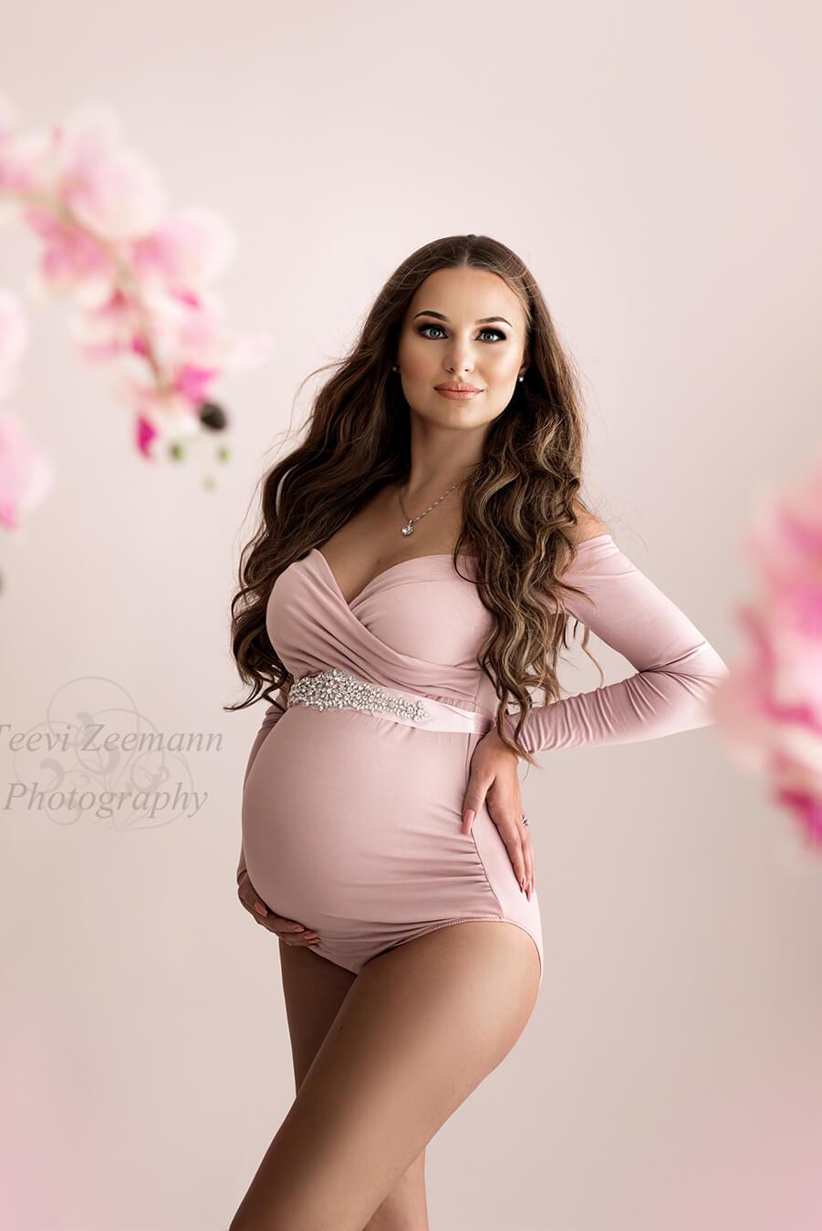 A pregnant woman is wearing a pink bodysuit. The bodysuit is off shoulder and has a sweetheart top. She is wearing a sash right under her breast